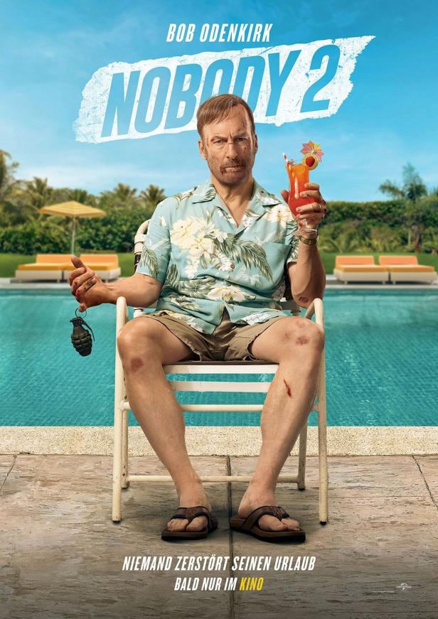 Bob Odenkirk sitzt mit einer Granate und einem Cocktail am Pool, im Hintergrund das Filmplakat „Nobody 2“.