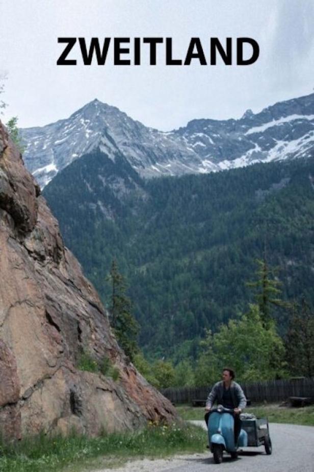 Ein Mann fährt mit einem blauen Motorroller und Anhänger vor einer Bergkulisse; darüber der Titel „Zweitland“.
