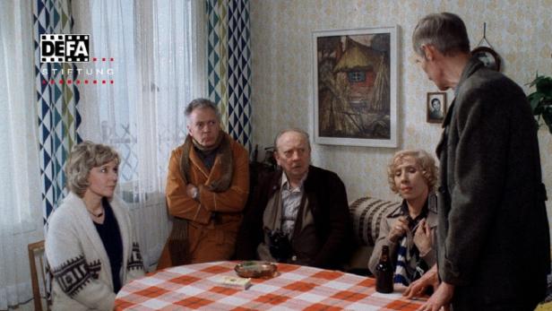 Fünf Personen sitzen an einem Tisch in einem Wohnzimmer.