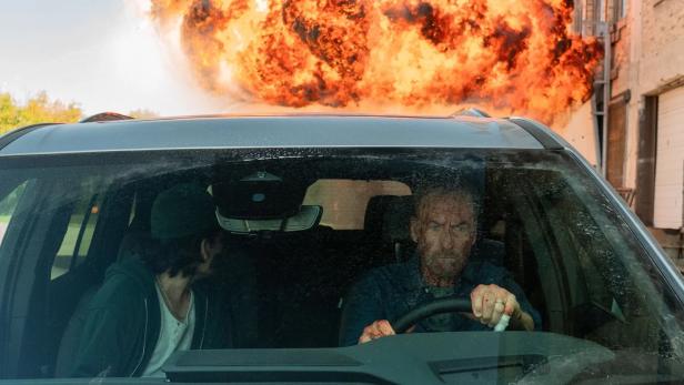 Bob Odenkirk fährt in „Nobody 2“ mit dem Auto davon, während hinter ihm feurige Explosionswolke in die Luft steigt.