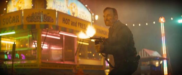 Bob Odenkirk feuert in „Nobody 2“ vor Freizeitparkkulisse aus einer Maschinenpistole.