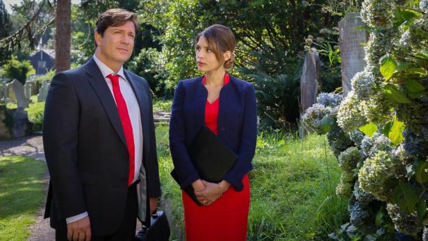 Ein Mann im Anzug und eine Frau im roten Kleid stehen auf einem Friedhof.