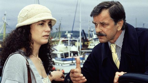 Eine Frau mit Hut und ein Mann mit Schnurrbart unterhalten sich vor einem Hafen.