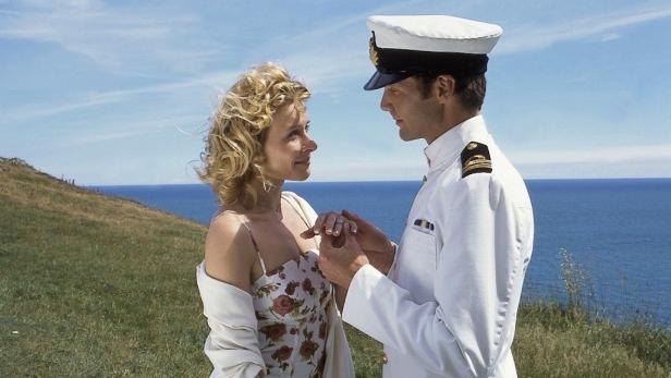 Ein Marineoffizier hält die Hand einer Frau vor einer Küstenlandschaft.