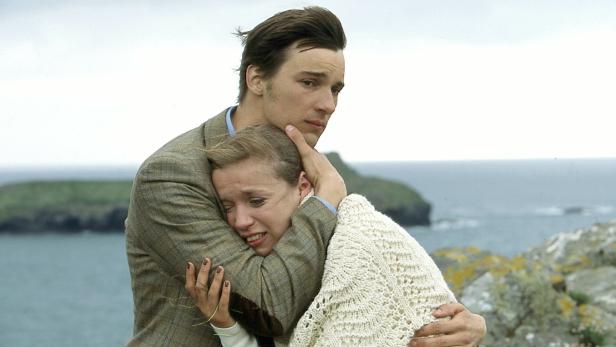 Ein Mann umarmt eine weinende Frau an einer Küste.