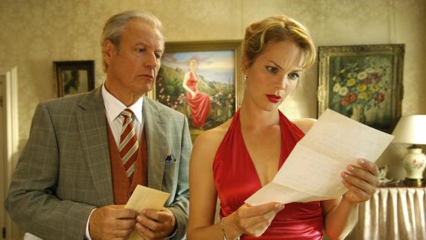 Eine Frau im roten Kleid liest einen Brief, während ein Mann im Anzug daneben steht.