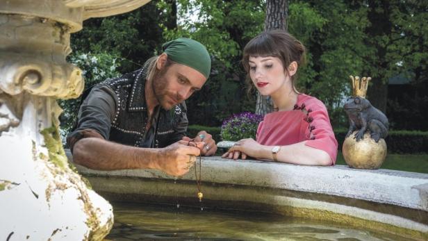 Ein Mann und eine Frau stehen an einem Brunnen, während der Mann einen goldenen Anhänger ins Wasser hält.