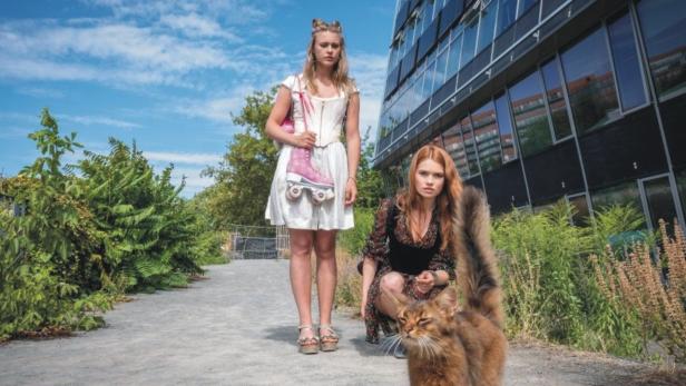 Zwei junge Frauen und eine Katze posieren vor einem modernen Gebäude. Eine trägt Rollschuhe über der Schulter.
