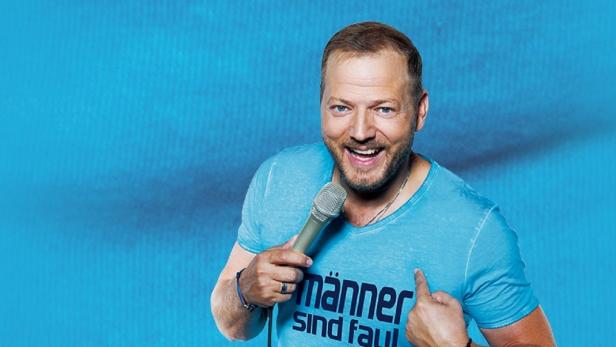 Ein Mann mit Mikrofon und einem T-Shirt mit der Aufschrift „Männer sind faul“ vor blauem Hintergrund.