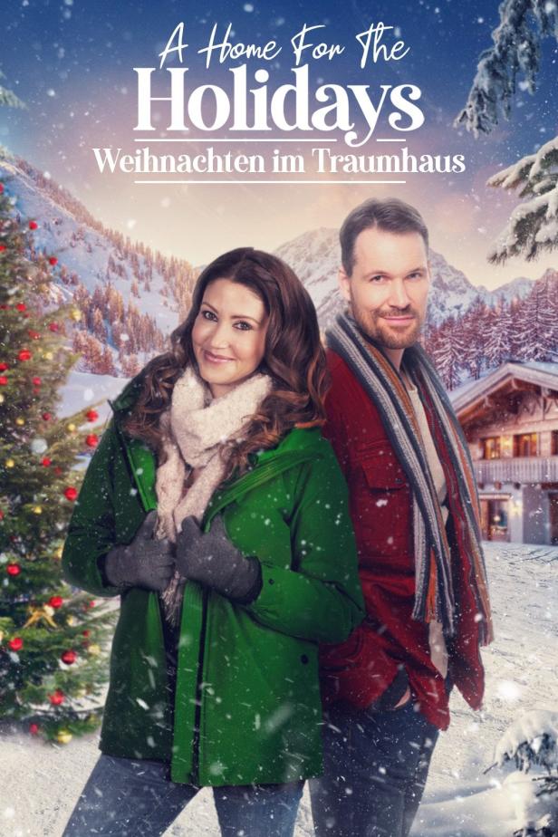 Ein Paar steht vor einem verschneiten Haus in den Bergen, wie im Film „Weihnachten im Traumhaus“.