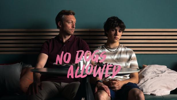 Ein Vater und sein Sohn sitzen an einem Tisch, im Vordergrund der Schriftzug „No Dogs Allowed“.