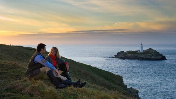 Ein Paar sitzt auf einer Klippe mit Blick auf einen Leuchtturm im Meer bei Sonnenuntergang.