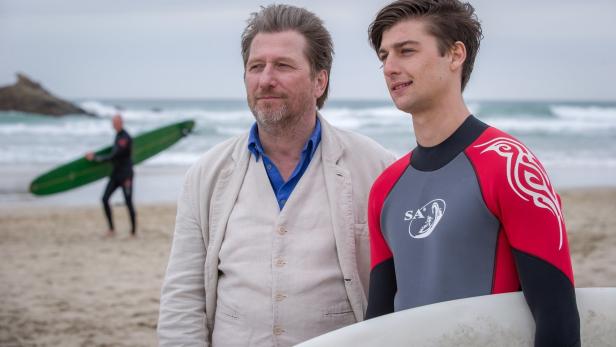 Zwei Männer stehen am Strand, einer im Anzug, der andere in einem Neoprenanzug mit Surfbrett.