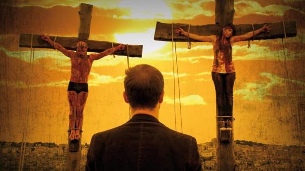 Ein Mann in Anzug blickt auf zwei gekreuzigte, blutverschmierte Personen vor einem Sonnenuntergang.