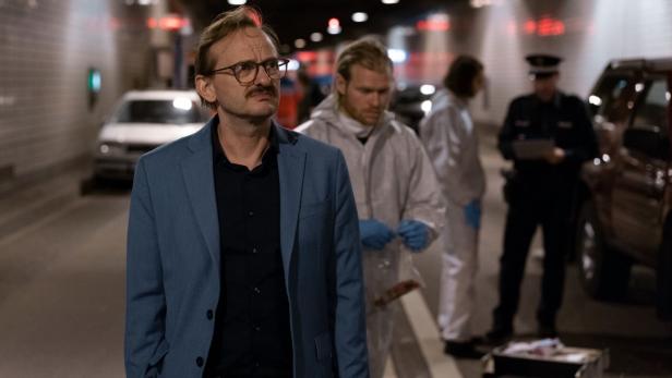 Ein Mann in einem blauen Sakko steht in einem Tunnel, im Hintergrund Spurensicherung und Polizei.