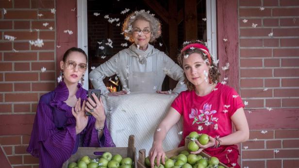 Drei Frauen posieren vor einer Holzkiste mit grünen Äpfeln, während Blütenblätter durch die Luft wirbeln.
