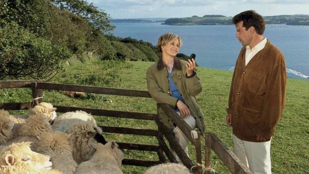 Eine Frau telefoniert auf einer Weide mit Schafen, während ein Mann daneben steht.