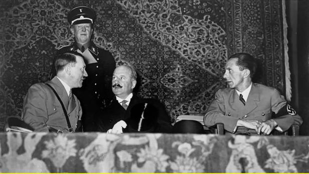 Adolf Hitler, Joseph Goebbels und weitere Personen sitzen an einem Tisch vor einem gemusterten Hintergrund.