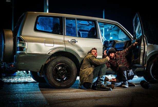 Zwei Männer kauern sich vor einen Geländewagen neben der offenen Fahrertür auf den Boden