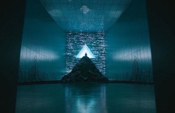 Futuristischr dunkler Raum  in dessem Hintergrund eine Person auf einer Art Erdhaufen steht  