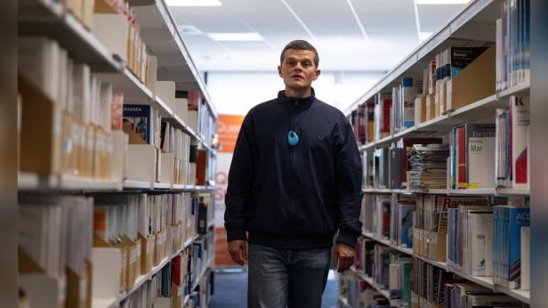 Ein Mann mit blauer Halskette steht zwischen Bücherregalen in einer Bibliothek.