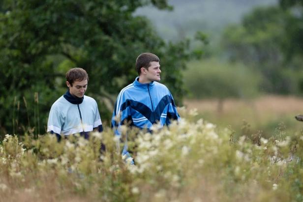 Zwei junge Männer in Trainingsjacken stehen auf einer Wiese mit Wildblumen, im Hintergrund Bäume und unscharfe Natur.