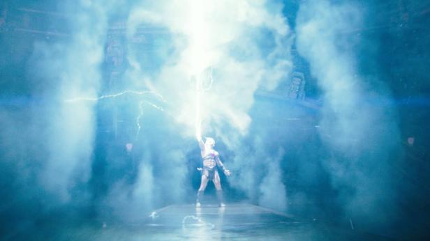 He-Man steht auf einer Bühne und lässt von seinem Schwert einen hellen Blitz in den blauen Nebel aufsteigen.