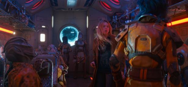 Eine blonde Frau steht in einem futuristischen Raumschiff-Innenraum inmitten von verschiedenen außerirdischen Wesen.