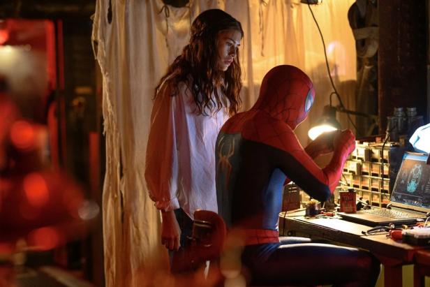 Spider-Man sitzt an einem Laptop und arbeitet an technischer Ausrüstung, während MJ daneben steht und ihm aufmerksam zusieht.