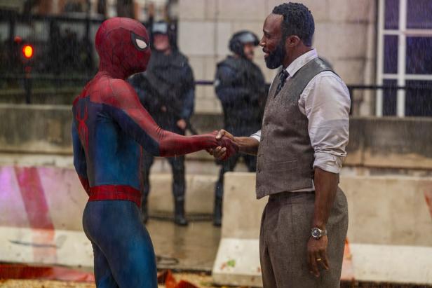Spider-Man schüttelt im Regen einem Mann in Weste und Hemd die Hand, während im Hintergrund Polizisten in Schutzausrüstung stehen.