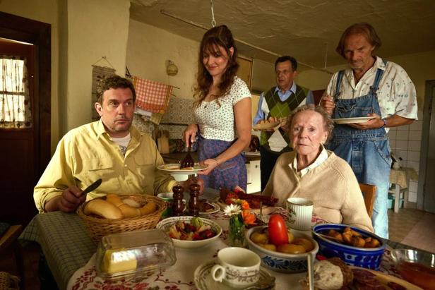 Mehrere Personen sitzen und stehen an einem reich gedeckten Tisch in einer Küche, während eine Frau Essen serviert.