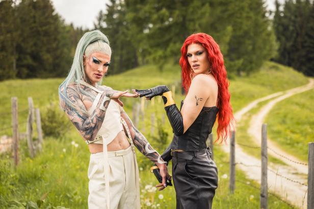 Zwei Dragqueens mit auffälligem Make-up und langen Haaren in Mintgrün und Rot posieren in einem ländlichen Gelände.
