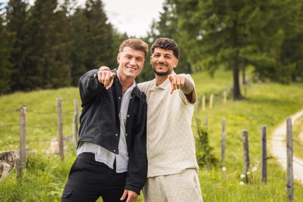 Zwei lächelnde Männer stehen auf einer Wiese und zeigen direkt in die Kamera.