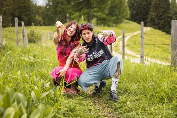 Zwei tätowierte Frauen posieren kauernd auf einem Wiesenweg und zeigen freche Handzeichen.
