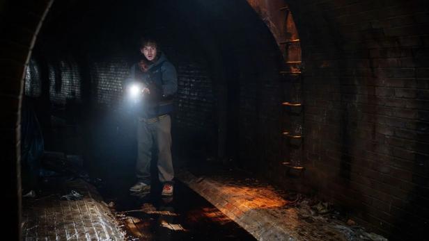 Person in dunklem Tunnel mit Taschenlampe, auf nassem Boden und vor rußigen Ziegelwänden.
