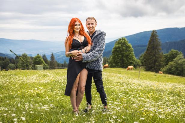 Ein Paar mit tätowierten Armen steht eng umschlungen auf einer blühenden Bergwiese vor einer alpinen Kulisse.