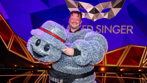 Ein lächelnder Mann hält vor dem „The Masked Singer“-Logo das Kostüm der „Bubble“ aus Luftpolsterfolie in den Händen.
