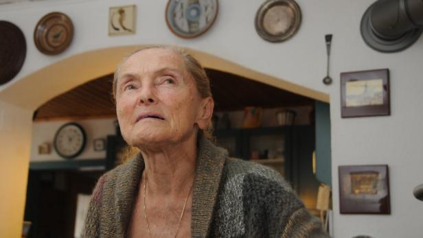 Erni Mangold mit nachdenklichem Blick trägt eine Strickjacke vor einer Wand mit dekorativen Tellern und Bildern.