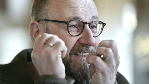 Ein Mann mit Brille und Bart lächelt und hält die Hände vor das Gesicht.