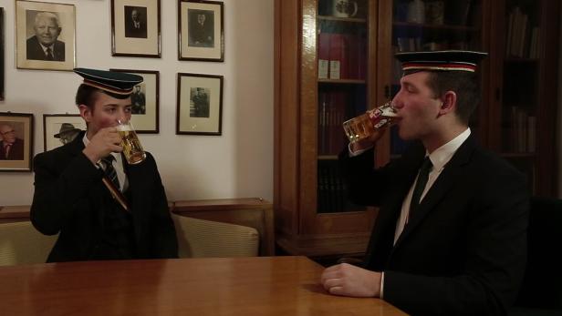 Zwei junge Männer in Anzügen und Studentenmützen trinken Bier an einem Tisch.