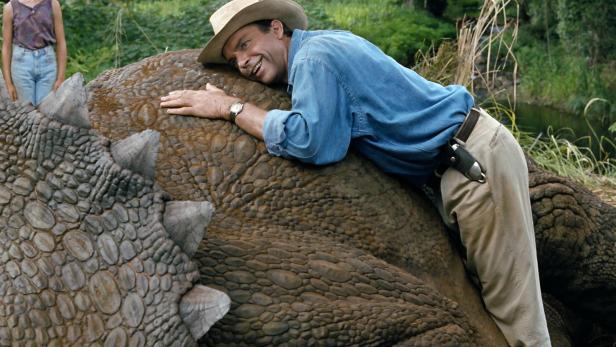 Ein Mann im Cowboyhut liegt lachend auf dem Rücken eines Triceratops.