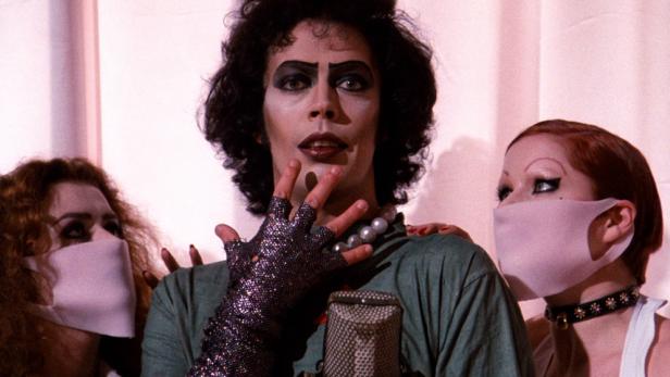 Frank N. Furter mit zwei maskierten Personen in einer Szene aus „The Rocky Horror Picture Show“.