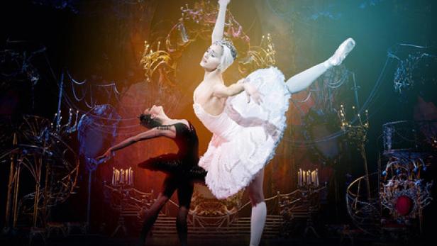 Zwei Ballerinen, eine in Weiß und eine in Schwarz, tanzen auf einer Bühne vor einem dunklen Hintergrund.