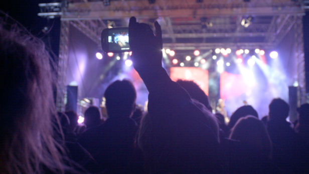 Eine Menschenmenge besucht ein Konzert, eine Person filmt die Bühne mit ihrem Smartphone.