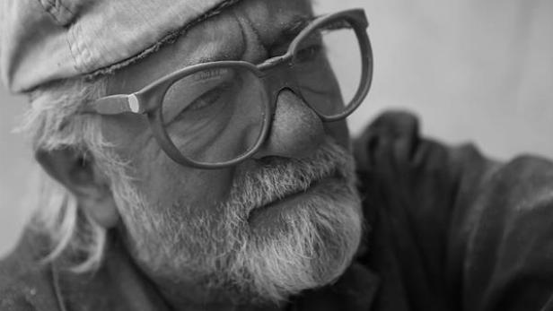 Ein älterer Mann mit Bart und Brille blickt nachdenklich zur Seite.