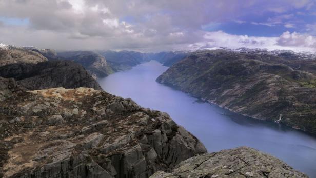 Blick von einem felsigen Gipfel auf einen Fjord, umgeben von Bergen unter einem bewölkten Himmel.