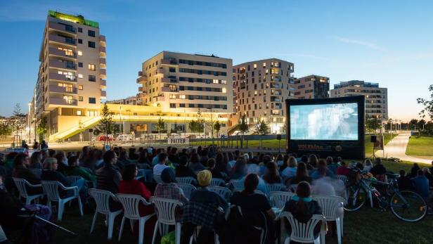 Zuschauer sitzen vor einer Leinwand bei einer Open-Air-Kinoveranstaltung in der Dämmerung.