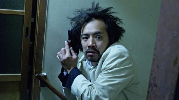 Ein Mann mit wirren Haaren zielt mit einer Pistole.