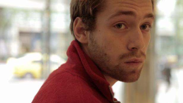 Ein junger Mann mit Bart und roter Jacke blickt über seine Schulter.