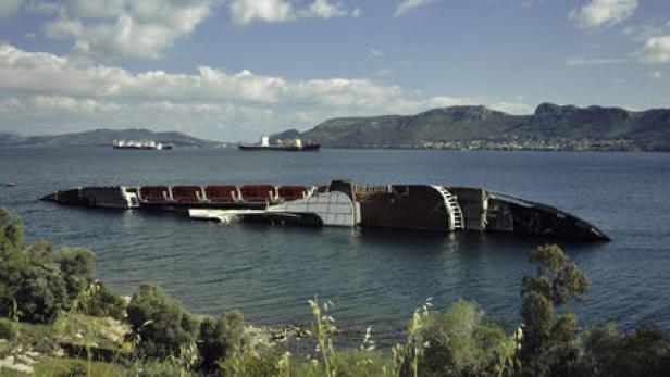 Ein gekentertes Schiff liegt im flachen Wasser vor einer bergigen Küstenlandschaft.